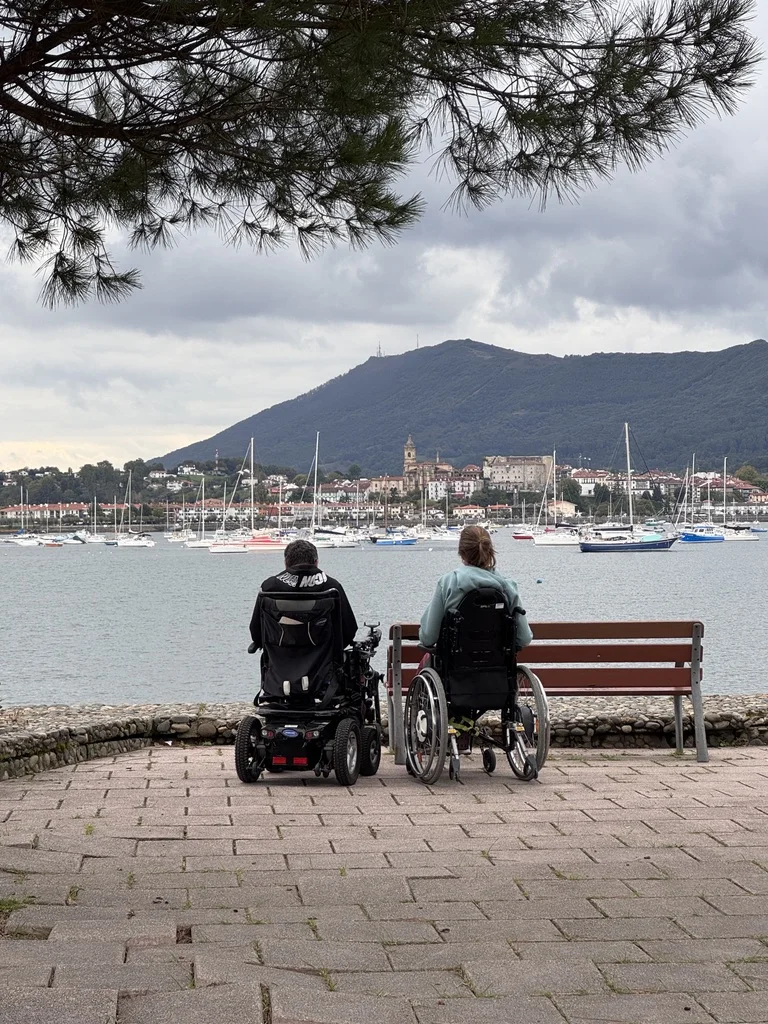 Two people in wheelchairs, seen from behind, facing the harbour of Hendaye and the mountains