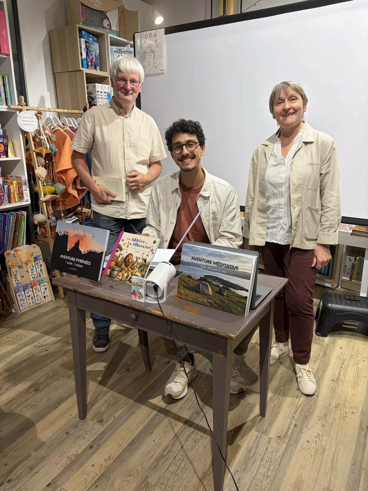Julien Porte between two booksellers at his signing table, with his books and a projector